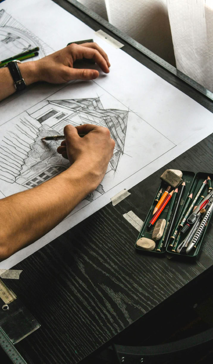 Person drawing architectural sketches on a drafting table with pencils and erasers nearby.